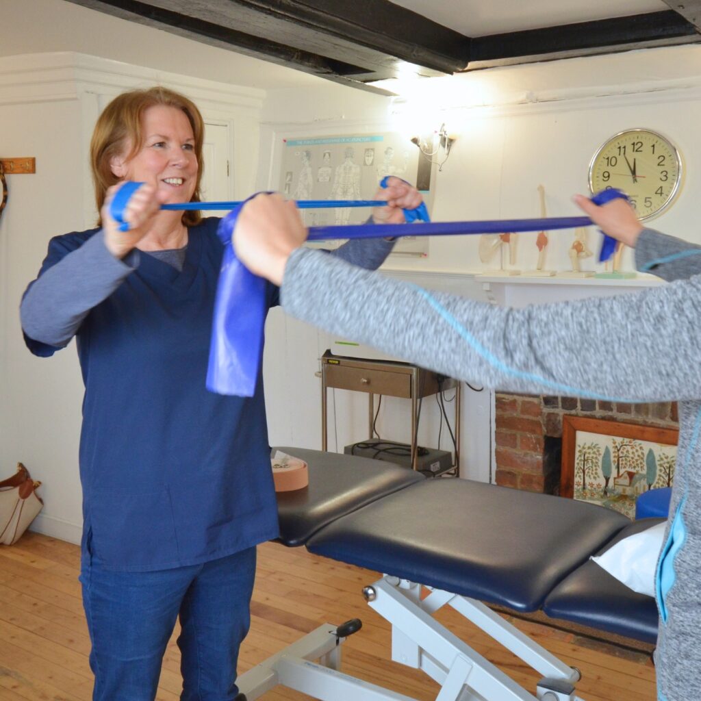 Physiotherapist demonstrating a strengthening exercise with a blue elastic band while patient follows from behind.